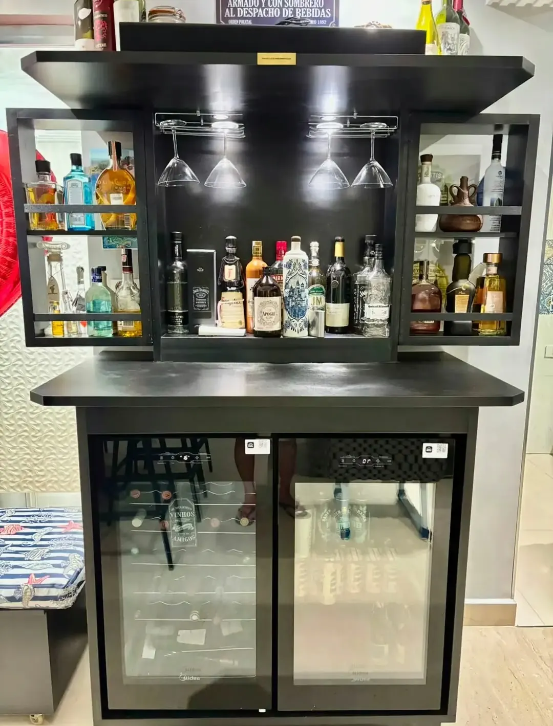 Bar preto com cristaleira iluminada, garrafas, taças penduradas e duas adegas embaixo