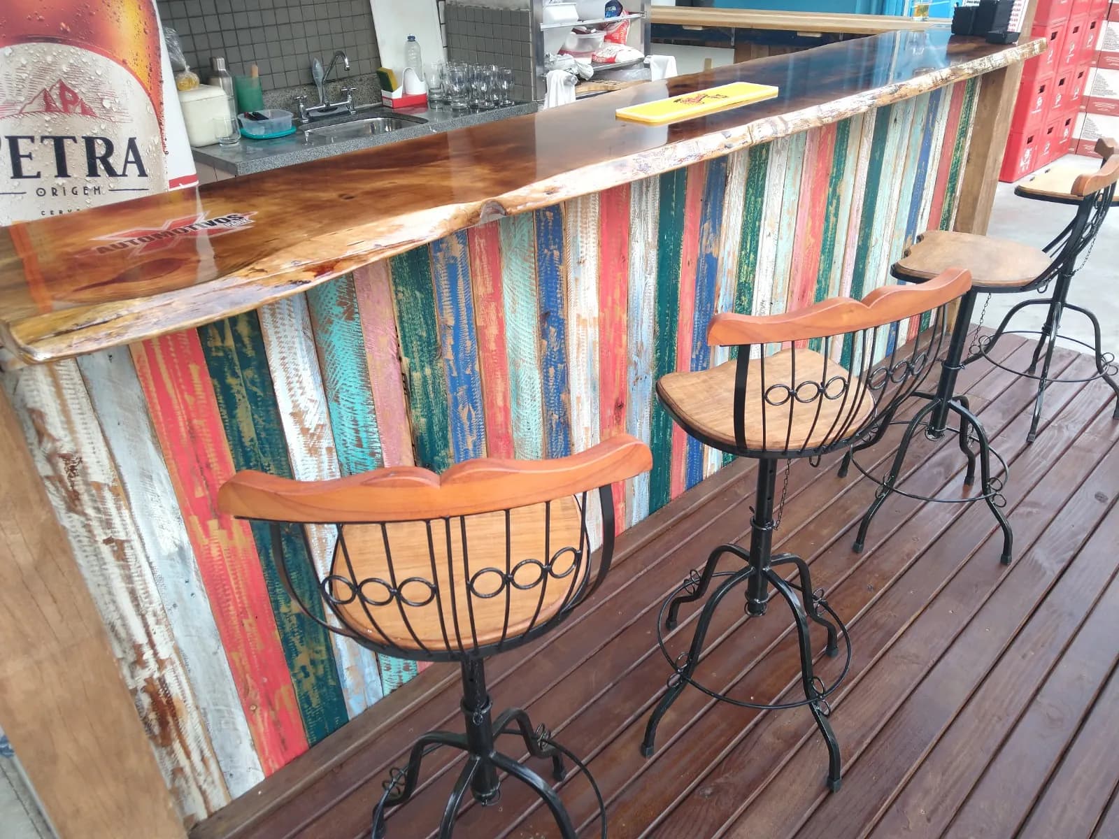 Balcão de bar com frente de ripas coloridas, tampo de madeira e banquetas sobre deck