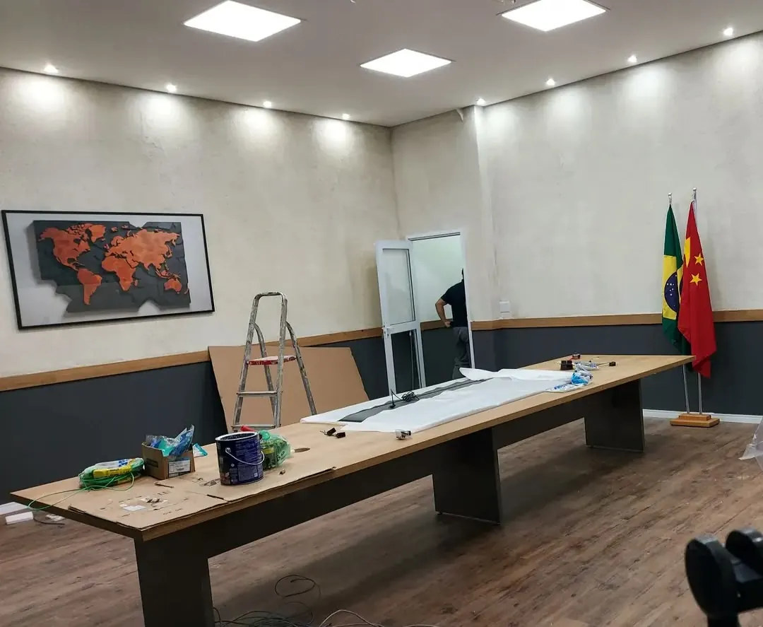 Mesa de reunião comprida com tampo de madeira, em sala de escritório com piso de madeira e bandeiras ao fundo